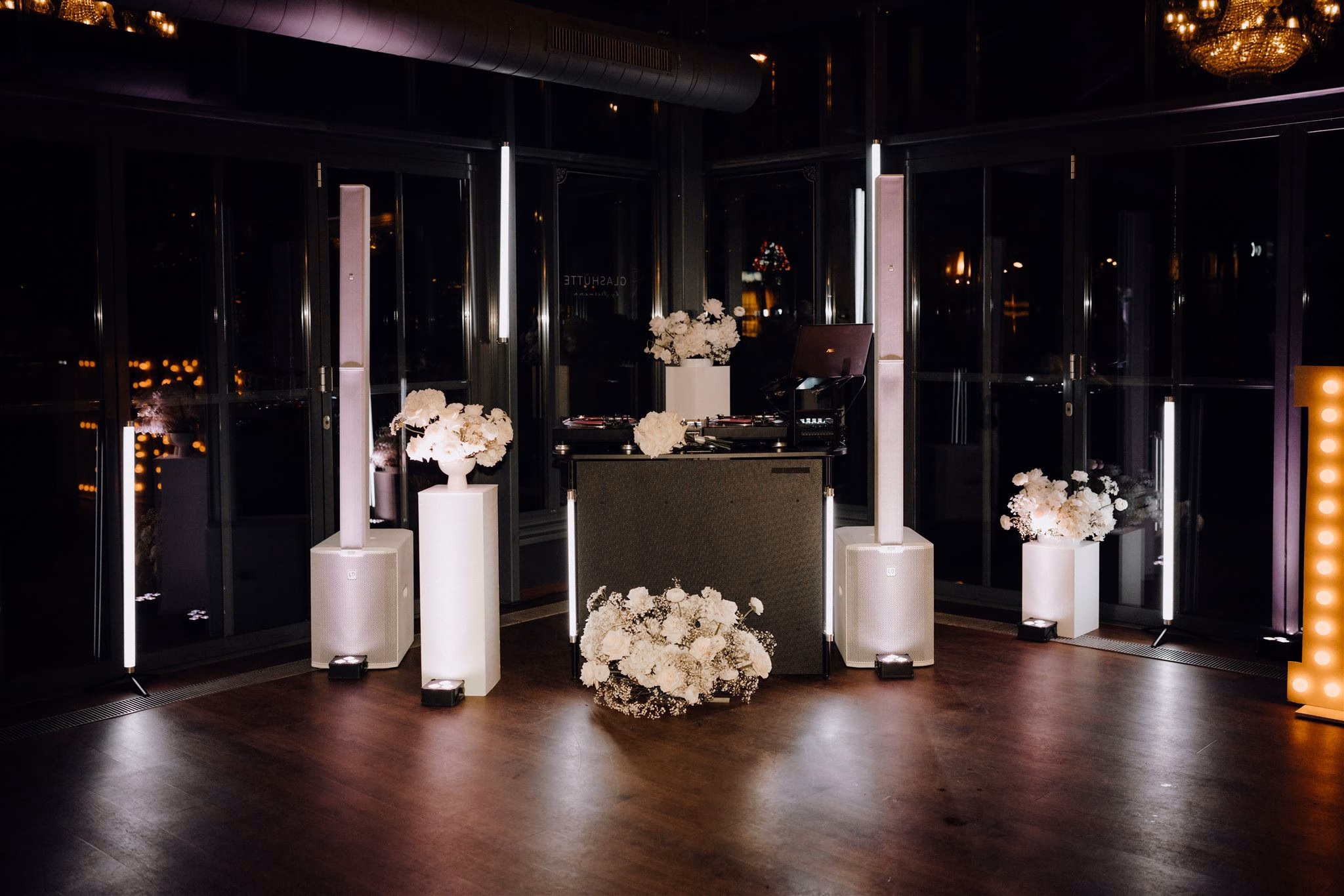 Ein DJ-Pult mit weißen Lautsprechern, Blumenarrangements und sanfte Beleuchtung bilden den Rahmen für eine Hochzeitsparty in einem dunklen, eleganten Raum - perfekt für eine Feier in Hannover oder Braunschweig.