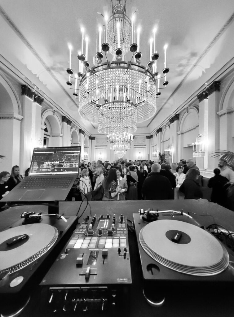 Ein DJ-Pult mit Plattenspielern überblickt einen überfüllten, eleganten Ballsaal unter großen Kronleuchtern.