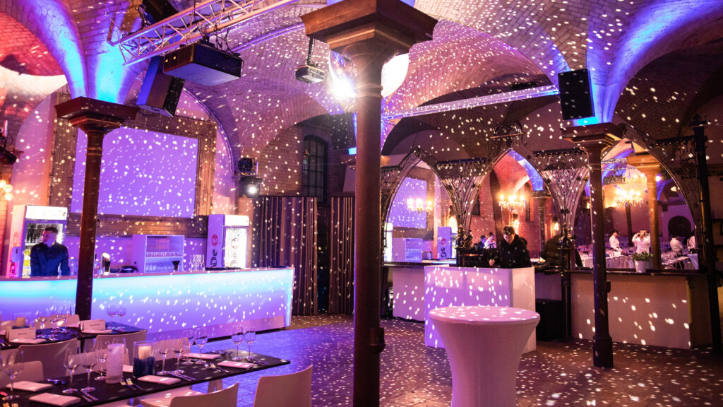 Ein stilvoller Veranstaltungsraum mit Discobeleuchtung, gewölbten Decken, gedeckten Tischen, beleuchteten Bars und Hochzeits-DJ.