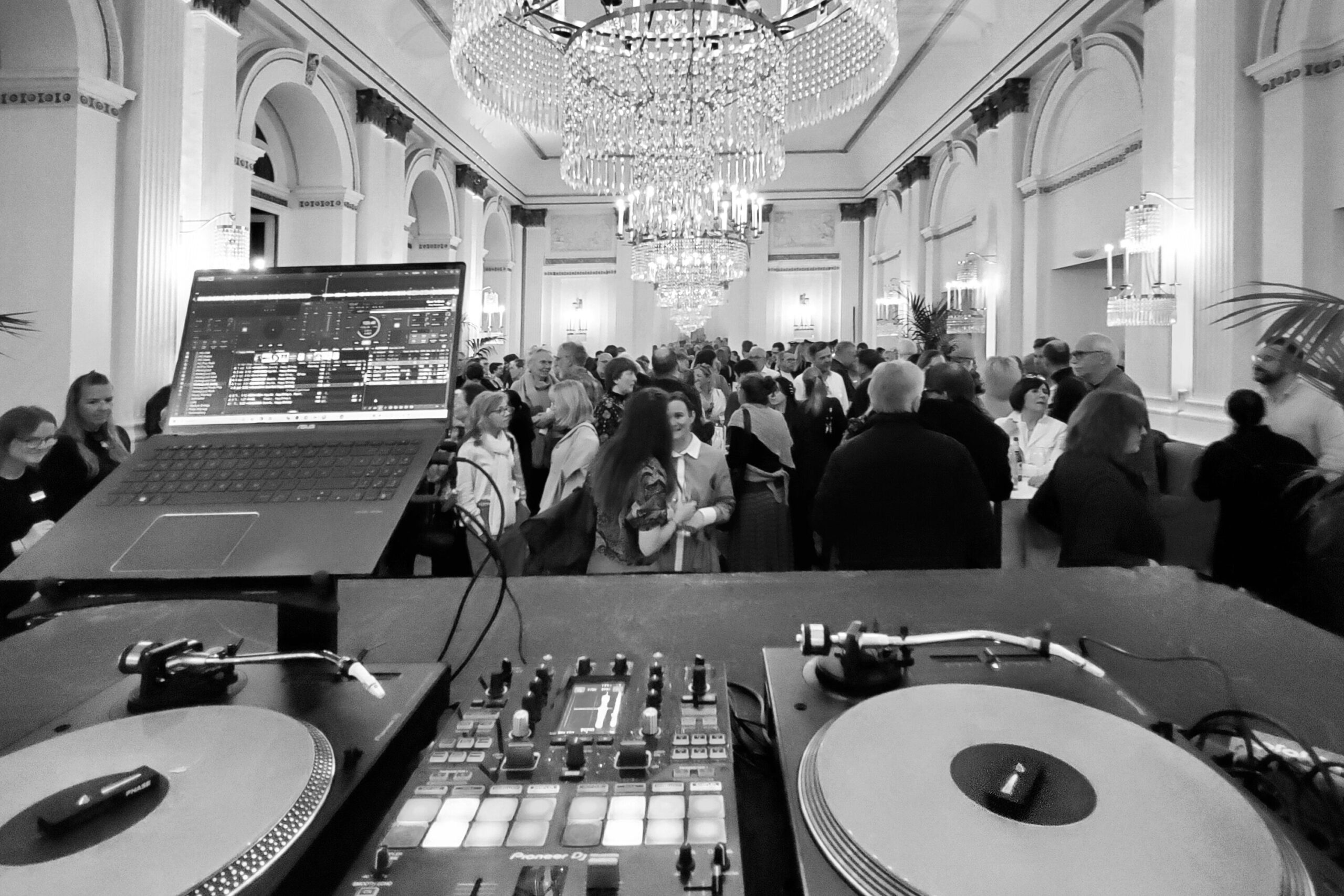 Ein Hochzeits-DJ-Pult mit Plattenspielern überblickt einen überfüllten, eleganten Ballsaal unter großen Kronleuchtern bei einer Hochzeitsfeier.