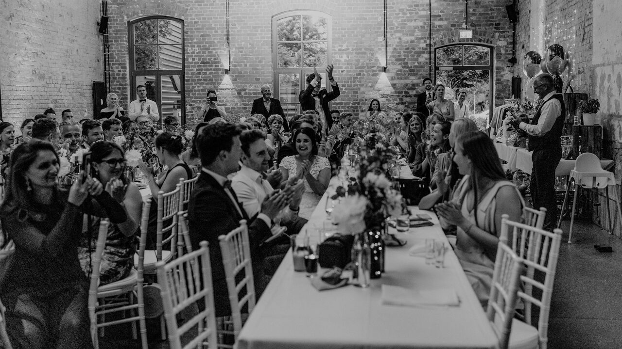 Gäste in formeller Kleidung applaudieren dem Hochzeits-DJ während eines Hochzeitsempfangs in einem rustikalen, gemauerten Lokal.