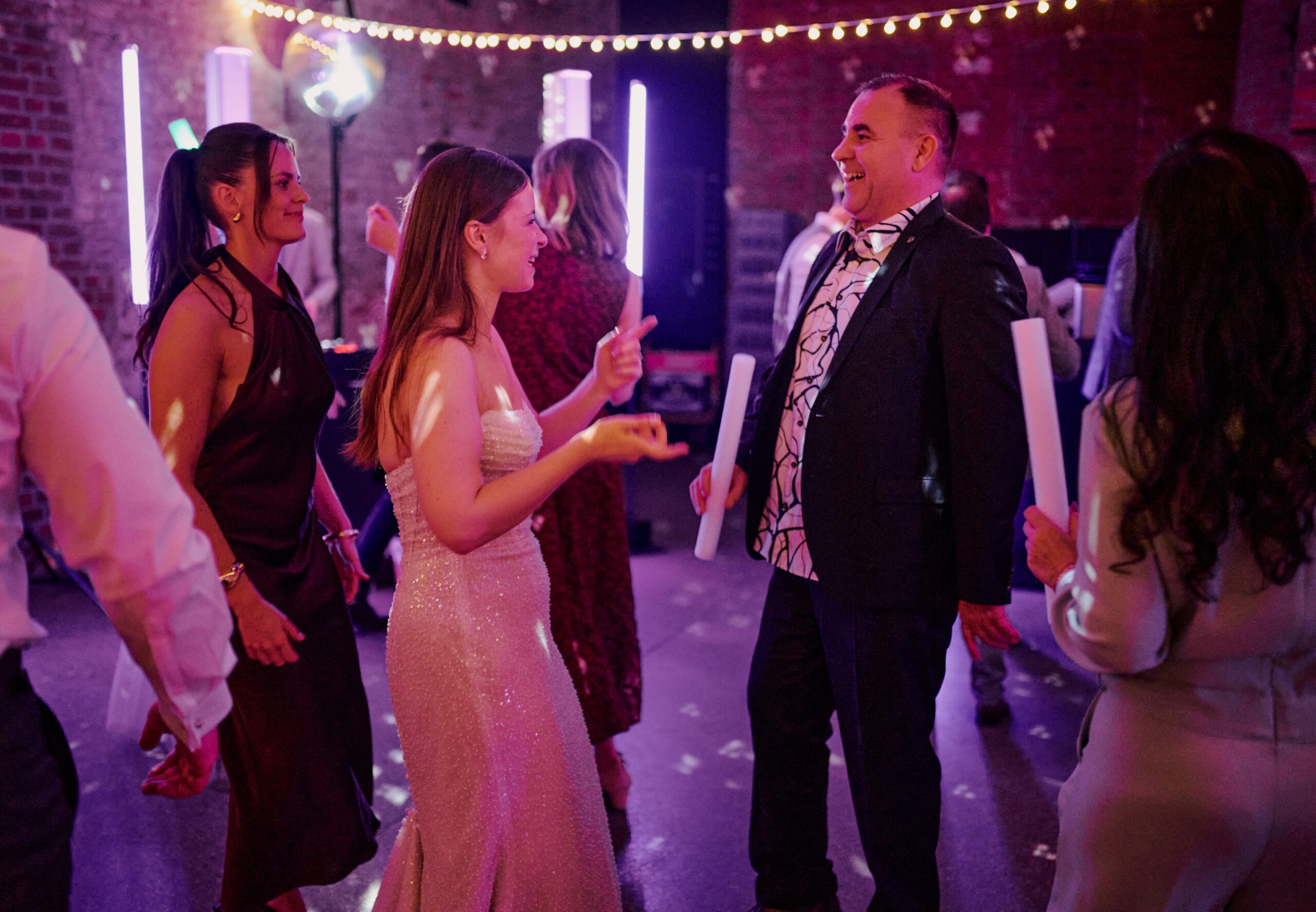 Menschen in formeller Kleidung tanzen und lächeln auf einer lebhaften Indoor-Party mit Lichterketten und Neon-Dekorationen.