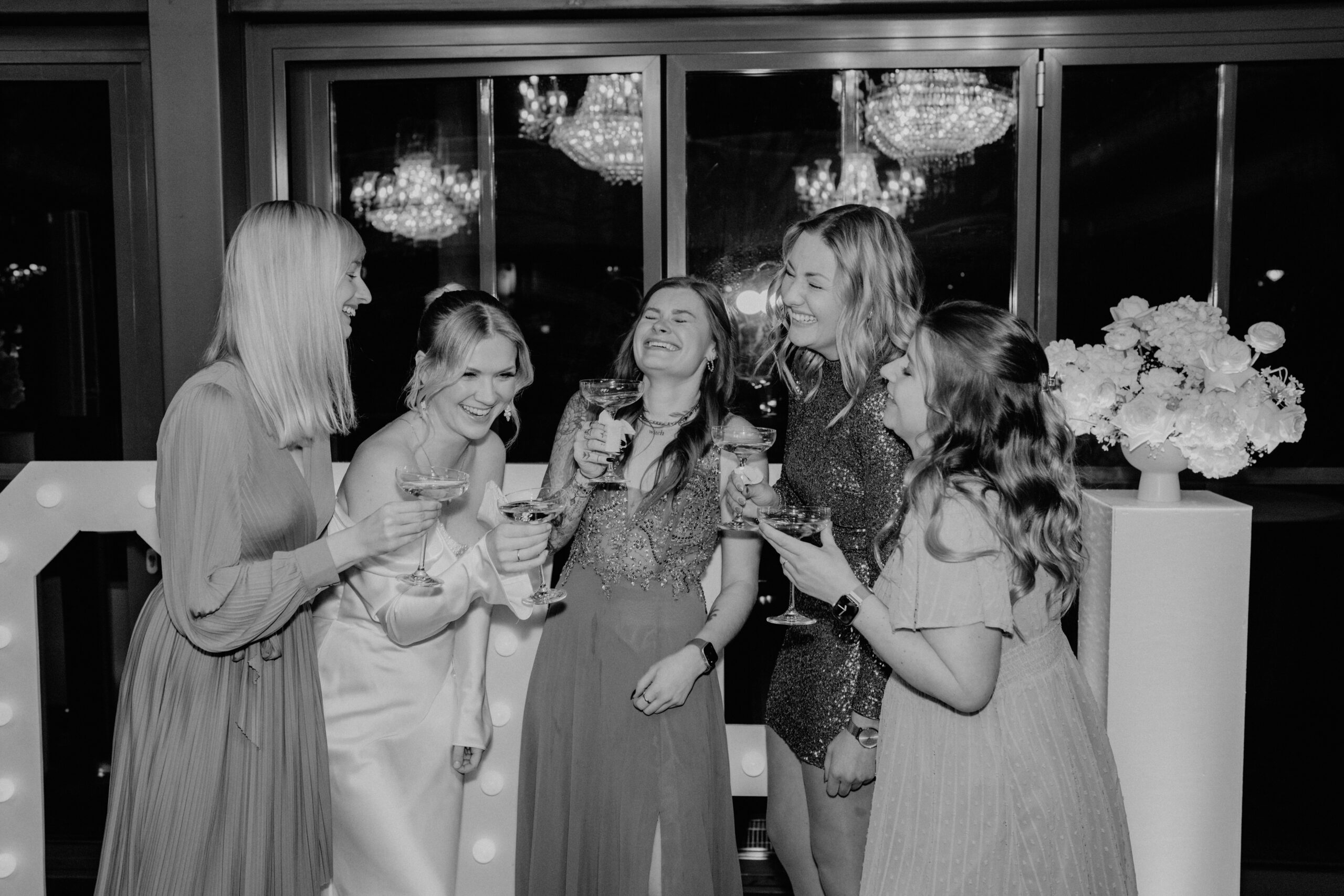 Fünf Frauen in Kleidern lachen und trinken gemeinsam auf einer Feier vor großen Fenstern und Kronleuchtern und genießen die ausgelassene Stimmung, die ein Top-Hochzeits-DJ aus Hannover erzeugt.