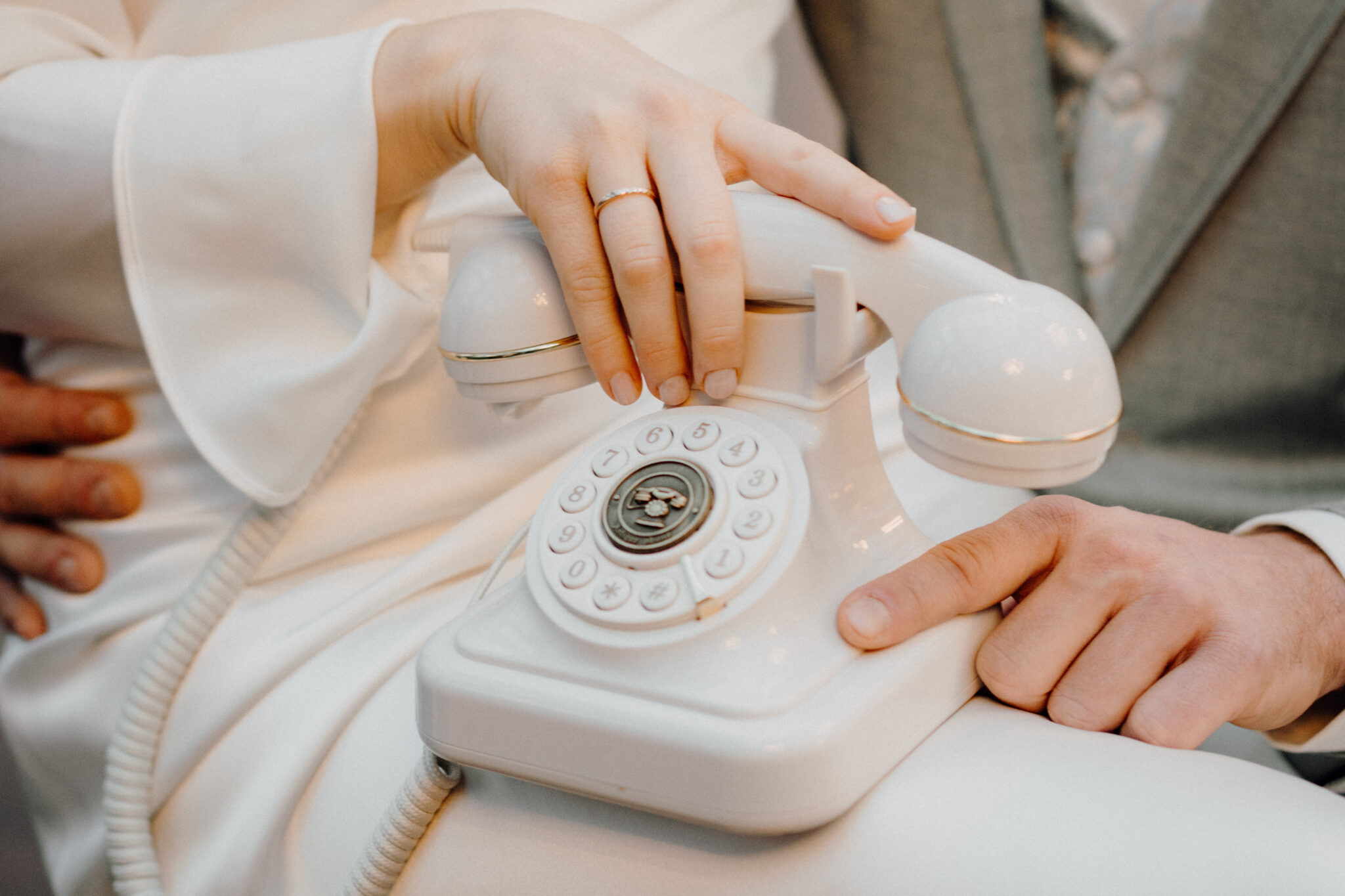 Eine Braut in einem weißen Kleid hält ein altes Telefon in der Hand, während sie auf dem Schoß des Bräutigams in einem grauen Anzug sitzt.