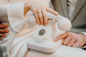 Eine Braut in einem weißen Kleid hält ein altes Telefon in der Hand, während sie auf dem Schoß des Bräutigams in einem grauen Anzug sitzt.