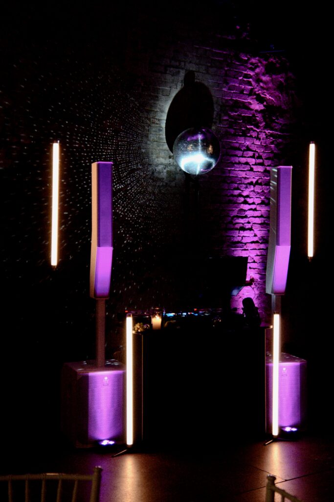 Ein DJ-Pult mit lila Lichtern, großen Lautsprechern und einer Discokugel vor einer Backsteinwand mit dramatischer Beleuchtung.